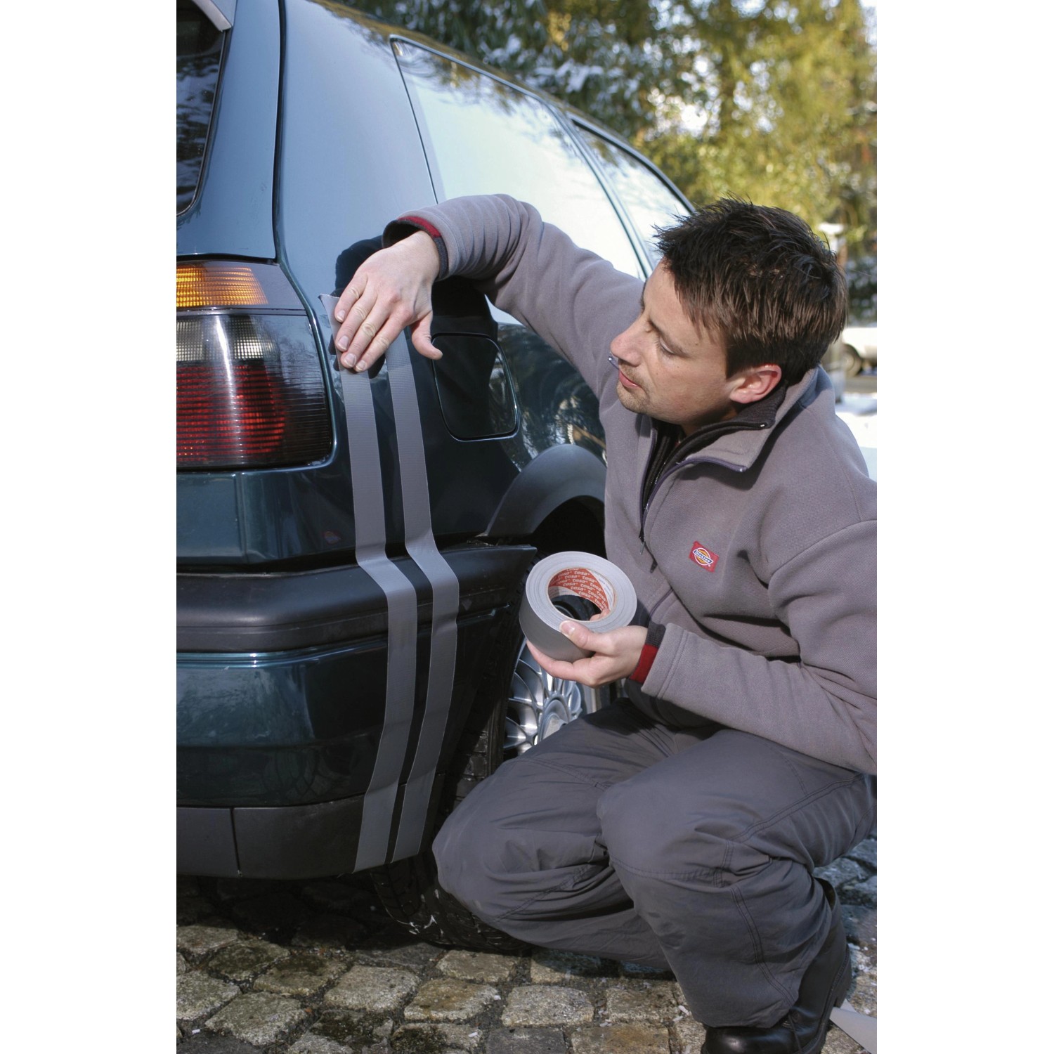 Mann klebt silbernes Tesa Extra Power Gewebeband auf ein Auto. Vielseitig einsetzbares Klebeband.