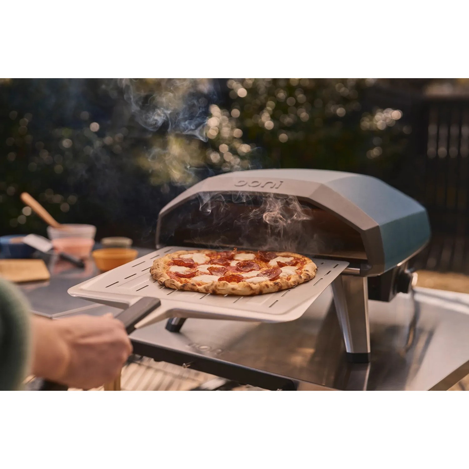 Ooni Koda 2 Gas-Pizzaofen in Schieferblau mit Pizza auf Pizzaschieber vor dem Ofen.