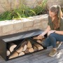 Schwarzes Westmann Kaminholzregal (104x34,5x40cm) mit Holzscheiten und einer Frau davor.