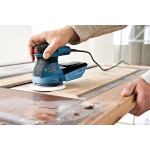 Person schleift Holz mit Bosch Professional Exzenterschleifer GEX 125-1 AE. Blaue Schleifmaschine auf Holzoberfläche.