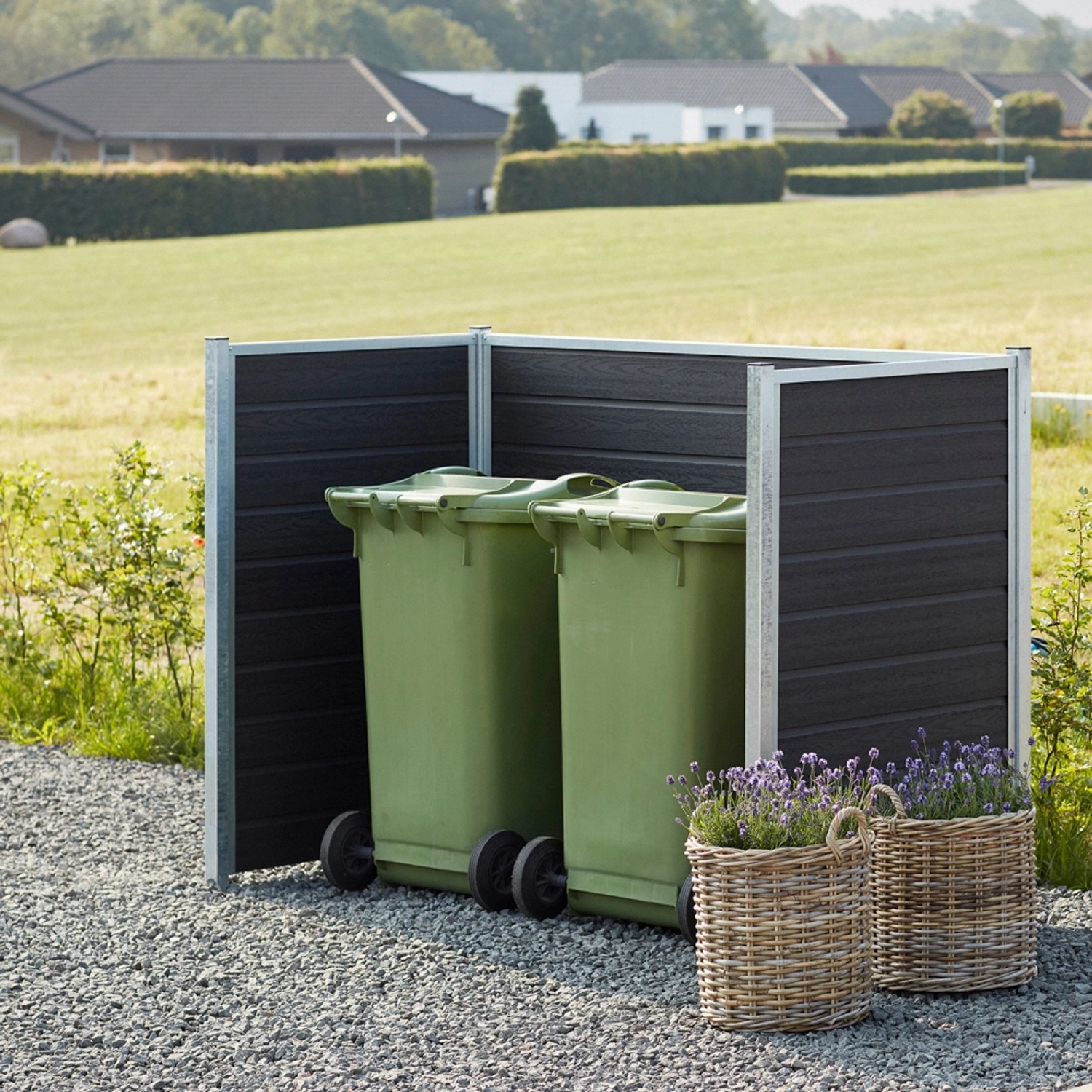 Artura Mülltonnenverkleidung aus WPC für 2 Tonnen, schiefergrau, mit Blumenkörben vor Kiesweg.