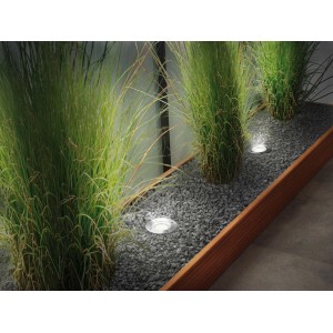 Runde Paulmann Plug & Shine RGBW Bodeneinbaustrahler im Gartenbeet mit grauen Steinen und Gräsern.