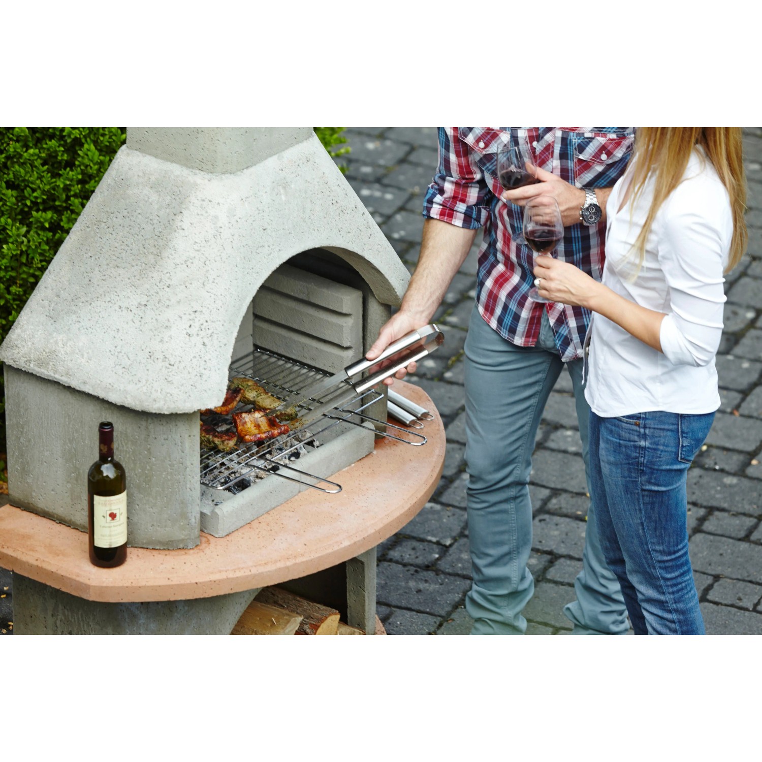 Buschbeck Grillkamin Carmen Grau-Terrakotta beim Grillen mit Paar im Garten.