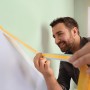 Mann klebt tesa® Malerband Perfect+ an eine Wand für scharfe Farbkanten.