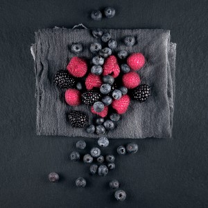 Glasbild Wild Berries, 20x20 cm: Himbeeren, Brombeeren und Blaubeeren auf grauem Stoff.