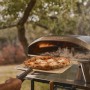 Ooni Koda 2 Pro Gas-Pizzaofen (2. Gen.) in Schwarzgrau mit Pizza auf Schaufel vor dem Ofen.