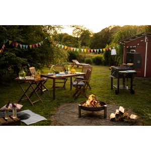 Gartenszene mit Greenville Klappstühlen aus Holz, Grill und Feuerstelle im Grünen.