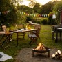 Gartenszene mit Greenville Klappstühlen aus Holz, Grill und Feuerstelle im Grünen.