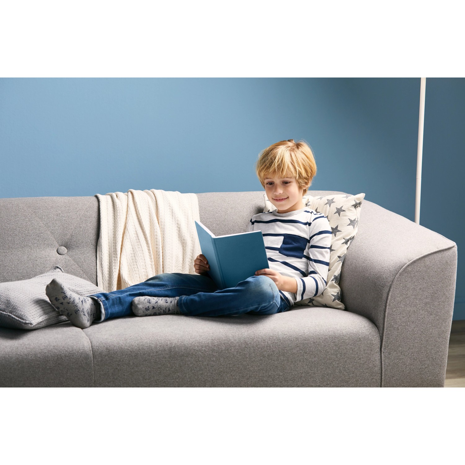 Junge liest Buch auf grauem Sofa mit Kissen und Decke vor blauer Wand.