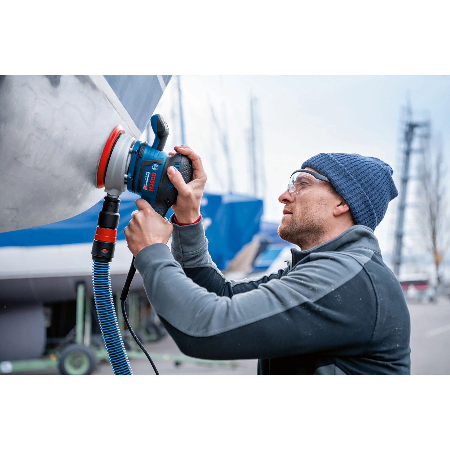 Mann schleift Boot mit Bosch Professional Exzenterschleifer GEX 40-150. Blaue Schleifmaschine mit Schleifteller und Absaugung.