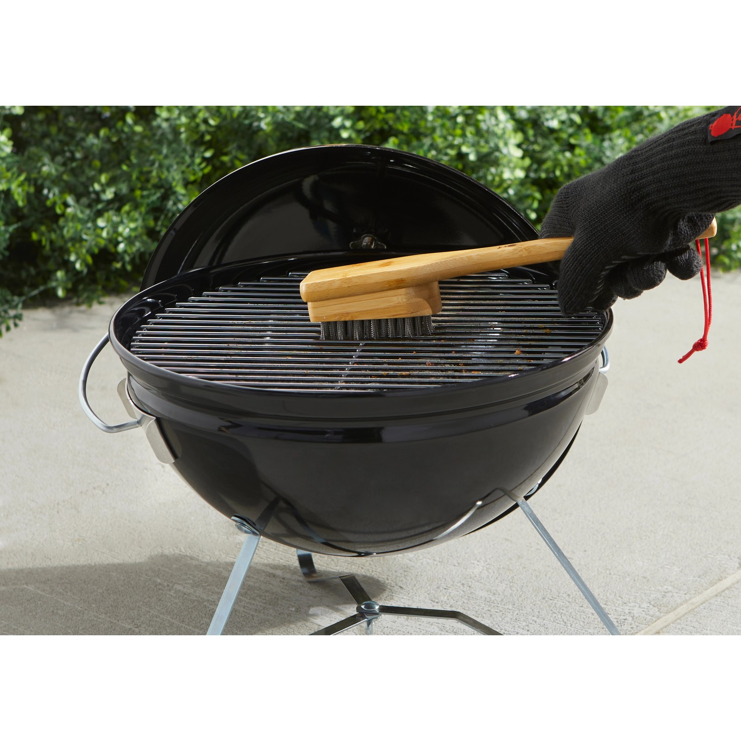 Weber Grillbürste mit Bambusgriff reinigt Grillrost. Holzkohlegrill im Hintergrund.