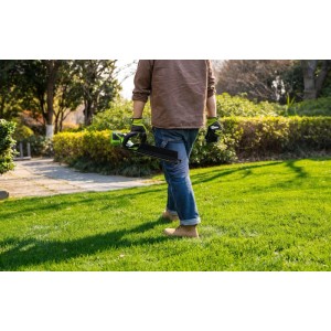 Gärtner trägt Greenworks Akku-Heckenschere mit 56 cm Klinge auf grünem Rasen. Gartenpflege mit Akku-System.