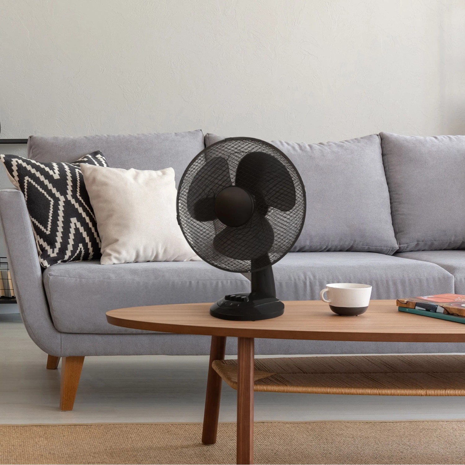 Schwarzer Tischventilator (Ø 30 cm) auf Tisch vor grauem Sofa. Ventilator mit Metallschutzgitter.