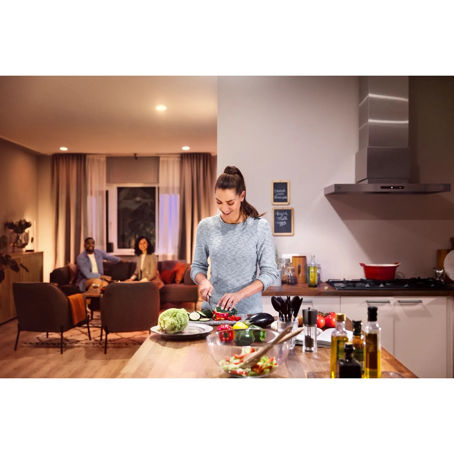 Szene mit Philips Hue E27 Lampe: Frau schneidet Gemüse in Küche, Wohnbereich mit warmem Licht im Hintergrund.