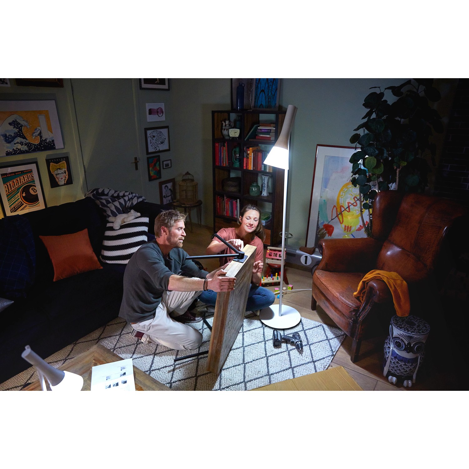 Szene mit Philips Hue Lampe: Paar montiert Bild im Wohnzimmer, beleuchtet von der smarten LED-Lampe.