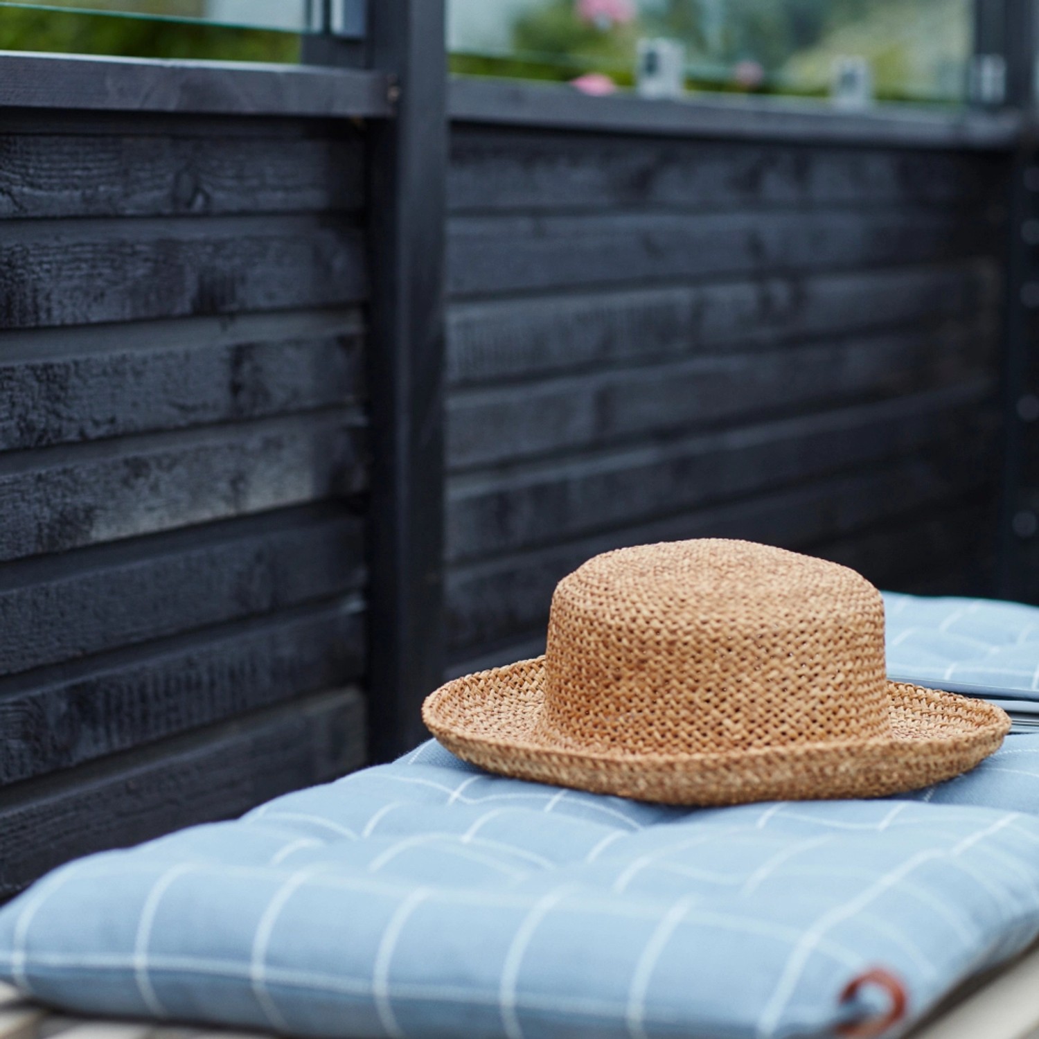 Schwarzer Sichtschutzzaun aus Nadelholz mit Glaselement und Strohhut auf blau kariertem Kissen.