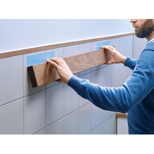 Mann befestigt Holzleiste mit Tesa Montageband an Fliesenwand. Starker Halt für dauerhafte Anwendungen.
