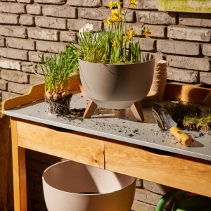 Grauer Siena Garden Pflanztopf Oslo (Ø 29 cm) mit Holzfüßen und Bepflanzung auf einem Gartentisch.
