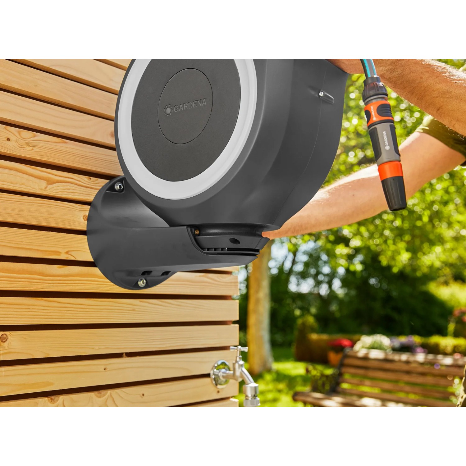 Gardena Wand-Schlauchbox RollUp S mit 15m Schlauch an Holzwand, mit Gartenschlauch und Spritze in der Hand.