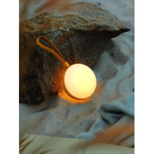 Nordlux Bring Akkuleuchte, weiß-gelb, Ø 13cm, mit gelbem Trageband, auf Sand vor Stein.