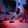 Schwarze Philips Hue Go Tischleuchte mit farbigem Licht auf einem Tisch mit Personen.