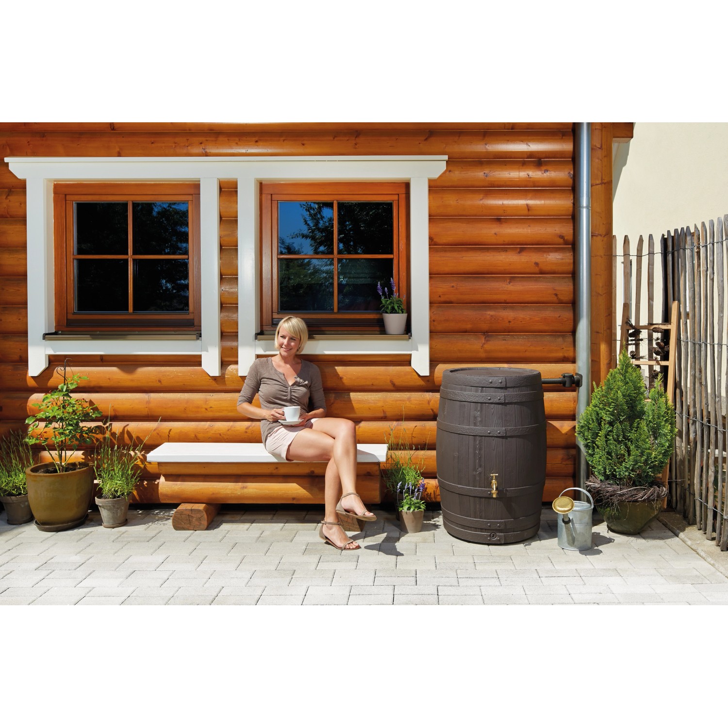 Garantia Regentonne Barrica 260 l in Dunkelbraun vor Holzhaus mit Frau auf Bank.