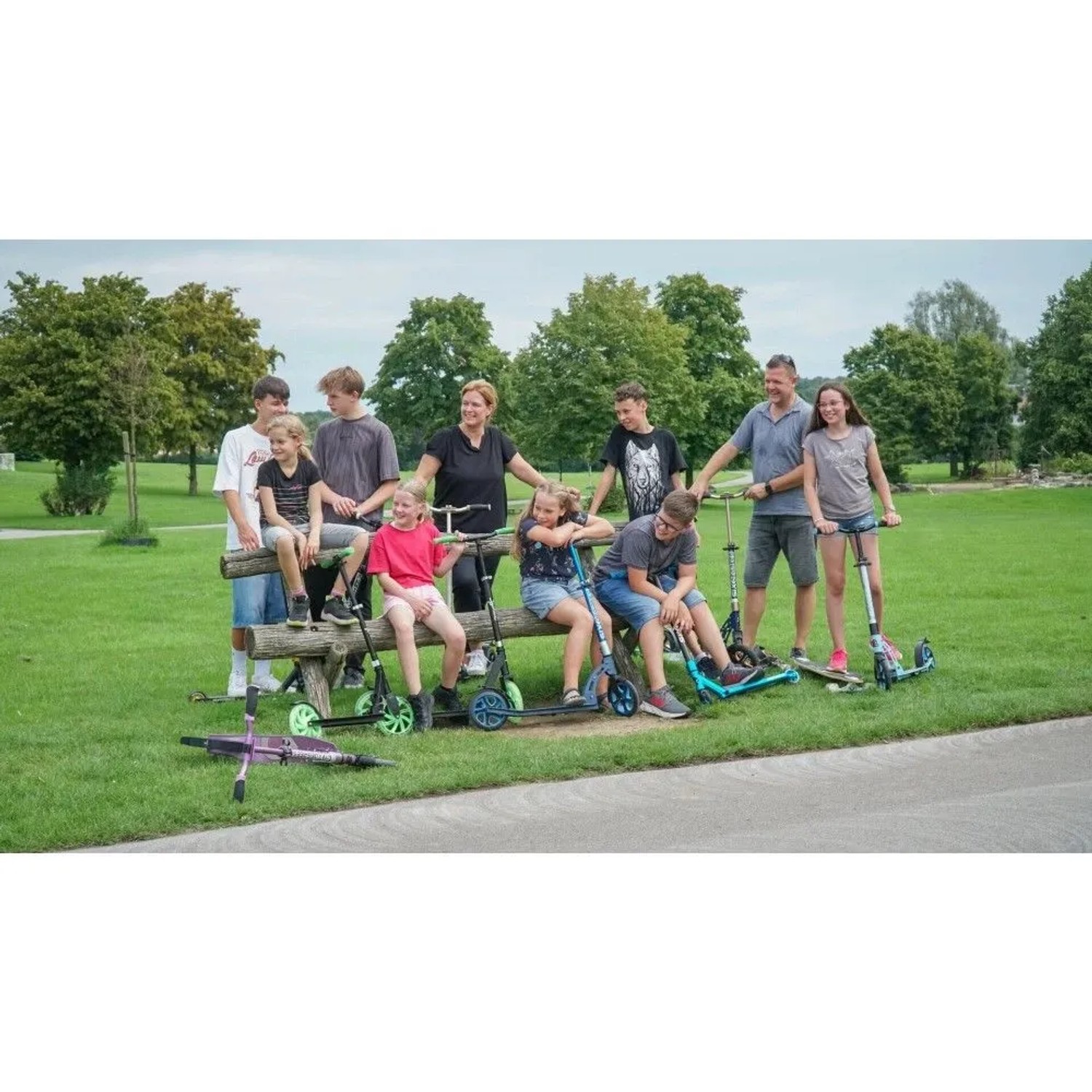 Gruppe von Erwachsenen und Kindern mit verschiedenen Six Degrees Scootern im Park. Ein türkiser Aluminium Scooter mit Klappmechanismus ist dabei.