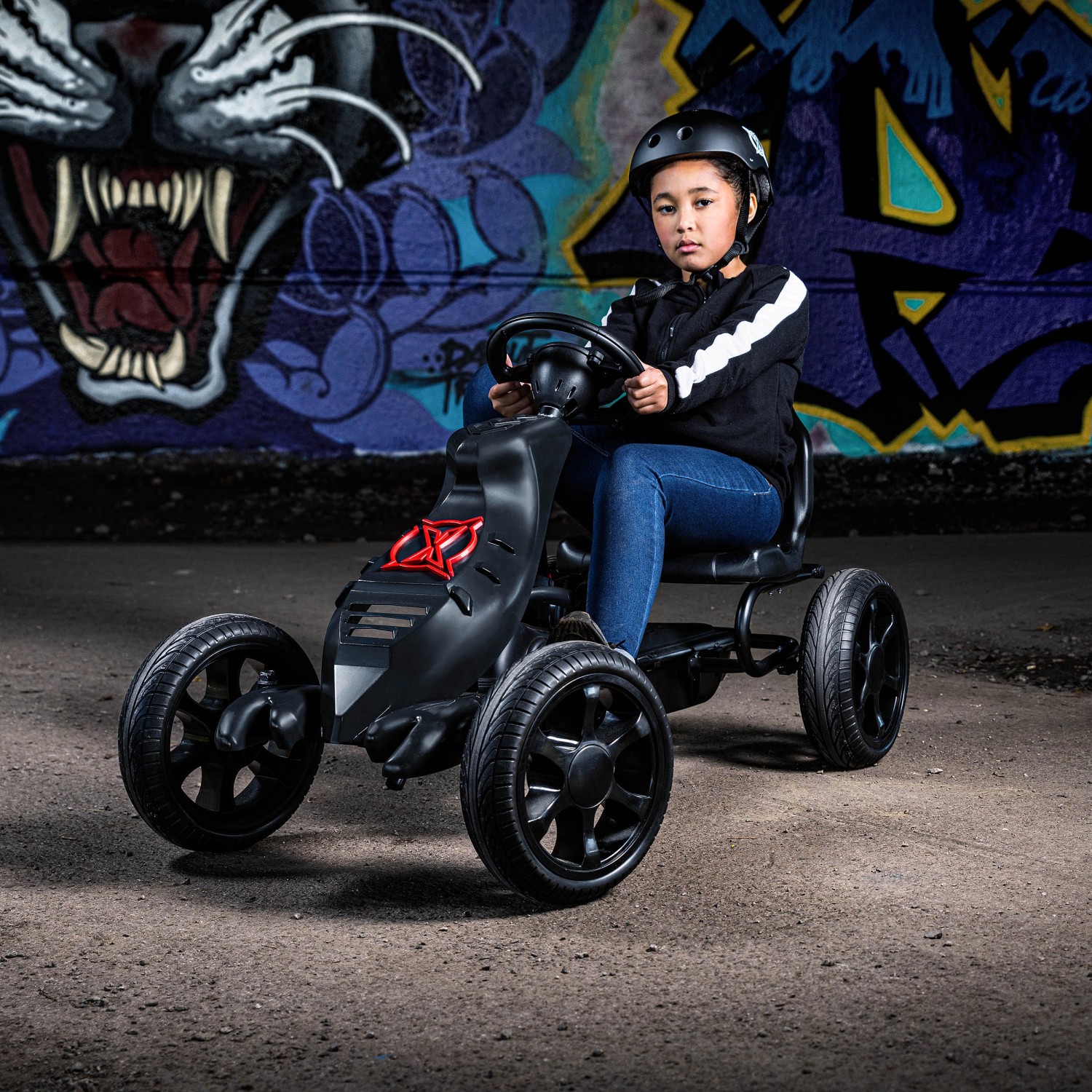 Schwarzes Xootz Go-Kart Venom mit Kind auf Betonboden vor Graffiti-Wand.