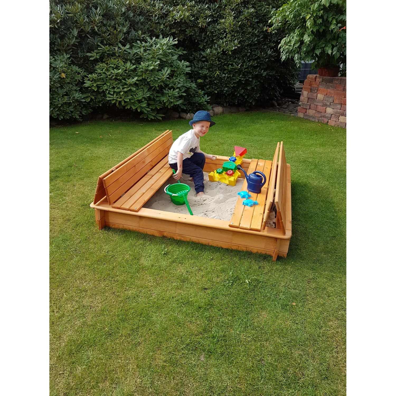 Holz Sandkasten Donald 140x140 cm mit klappbarer Abdeckung als Sitzbank und spielendem Kind.