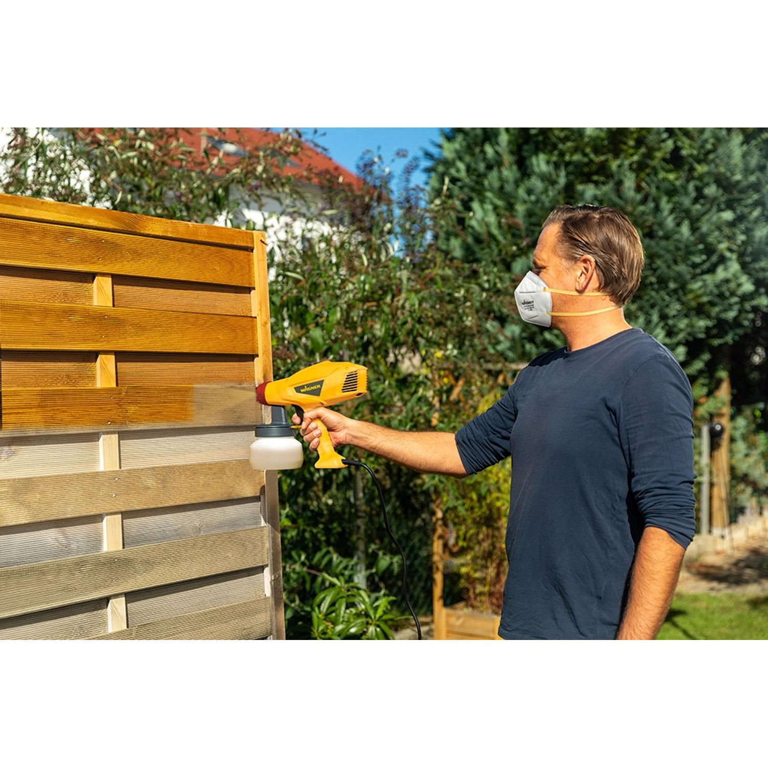 Mann sprüht mit Wagner Farbsprühsystem W 125 Holzschutz auf einen Zaun.
