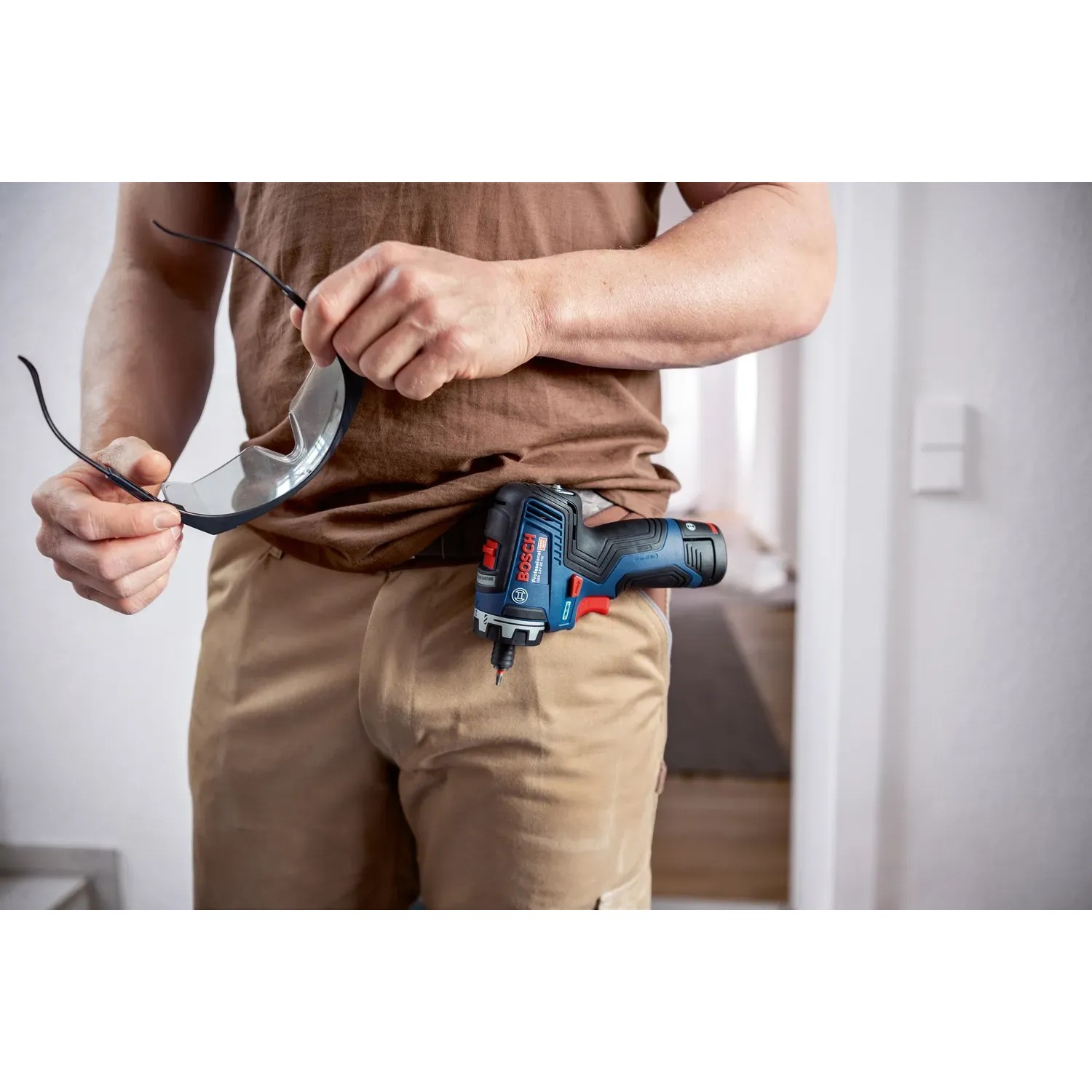 Bosch Professional GSR 12V-35 HX Akku-Bohrschrauber am Gürtel, mit Schutzbrille in der Hand.