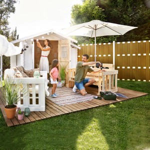 Familie baut sandfarbenen WPC Sichtschutzzaun im Garten mit Werkzeug und Gartenhaus.