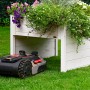 Weisse Mährobotergarage mit Pflanzkasten und Mähroboter auf Rasen.