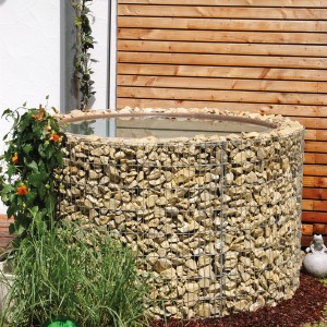 Gabionen-Wasserfass 750 l im Garten, befüllt mit hellen Steinen. Regentonne zur Gartenbewässerung.