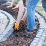 Person verteilt Rindenmulch (0-40 mm) im Gartenbeet mit Steineinfassung.