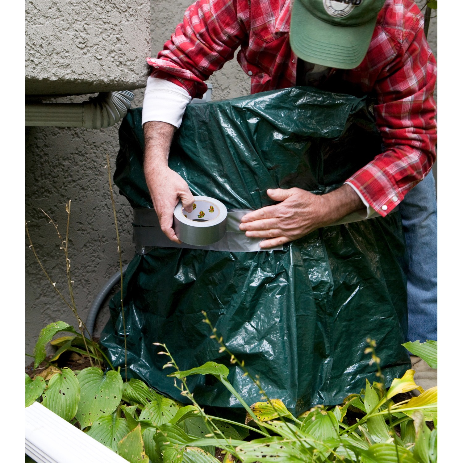 Mann fixiert grüne Plane mit Duck Tape Gewebe-Klebeband Silber an einer Außeneinheit.