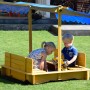 Dobar Sandkasten mit Dach, gelbes Holz, blaue Markise, zwei Kinder spielen im Garten.