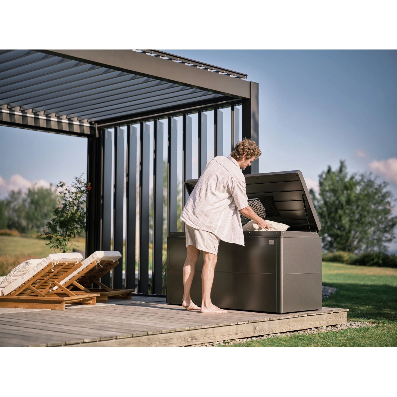 Dunkelgraue Biohort Loungebox 160 auf Holzterrasse, Mann verstaut Kissen. Regenwasserdichte Universalbox für Gartenzubehör.