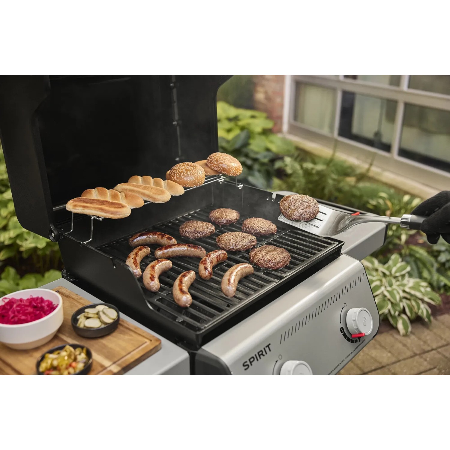 Weber Gasgrill E-210 Spirit 2025 in Schwarz mit Würstchen, Burger und Brötchen auf dem Grill.