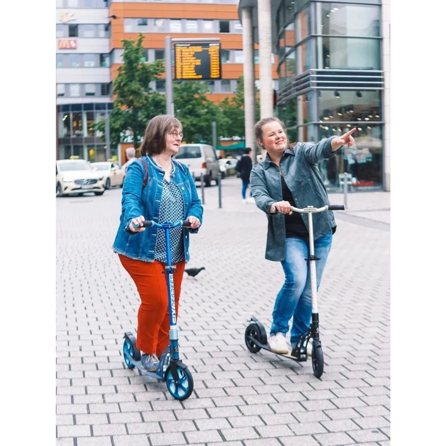 Zwei Frauen fahren auf Six Degrees Aluminium Scootern mit Luftbereifung in der Stadt. Blaues Modell mit Klappmechanismus, höhenverstellbar für Sport.
