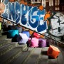 Mehrere farbige Sitting Point Hocker Scuba (60 l) auf einer Treppe vor Graffiti-Wand.