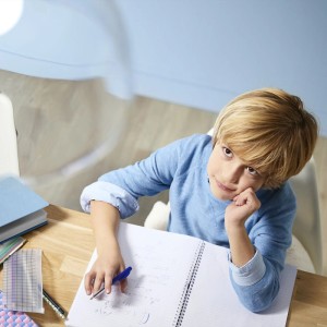 Junger blonder Junge in blauem Pullover sitzt am Schreibtisch, blickt nachdenklich nach oben, Notizbuch und Stift.