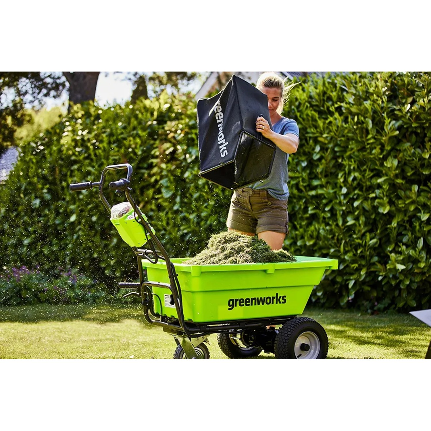 Grüne Greenworks Akku-Schubkarre G40GC mit Gras befüllt, Frau leert Fangkorb.
