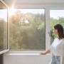 Frau am Fenster mit Tesa Pollenschutzgitter Anthrazit. Effektiver Schutz vor Pollen.