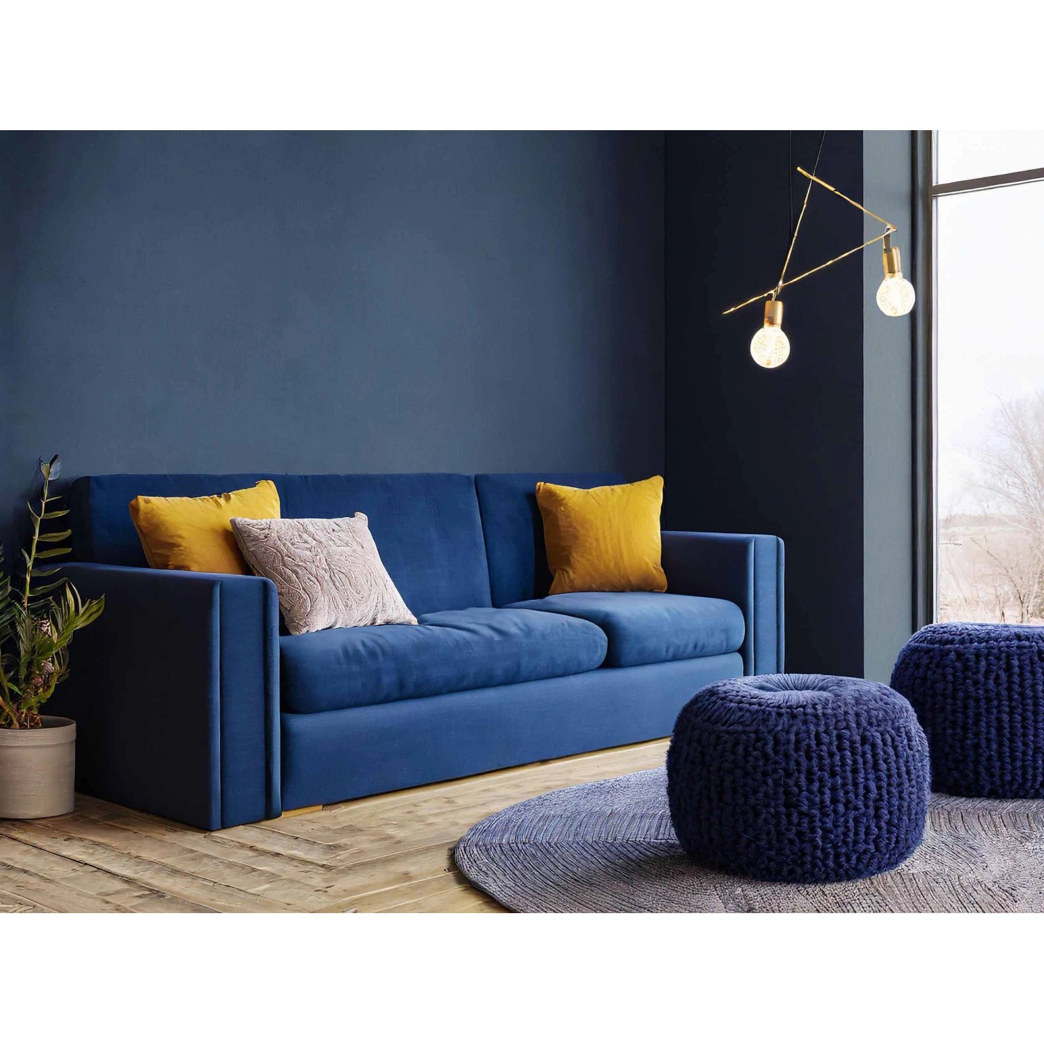 Blaues Sofa mit Kissen und Strickhockern vor blauer Wand.