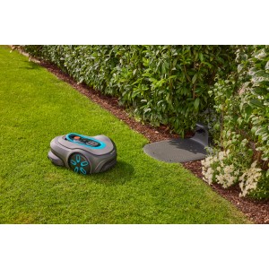 Gardena Smart Sileno Max Mähroboter mit Ladestation auf Rasenfläche vor Hecke.