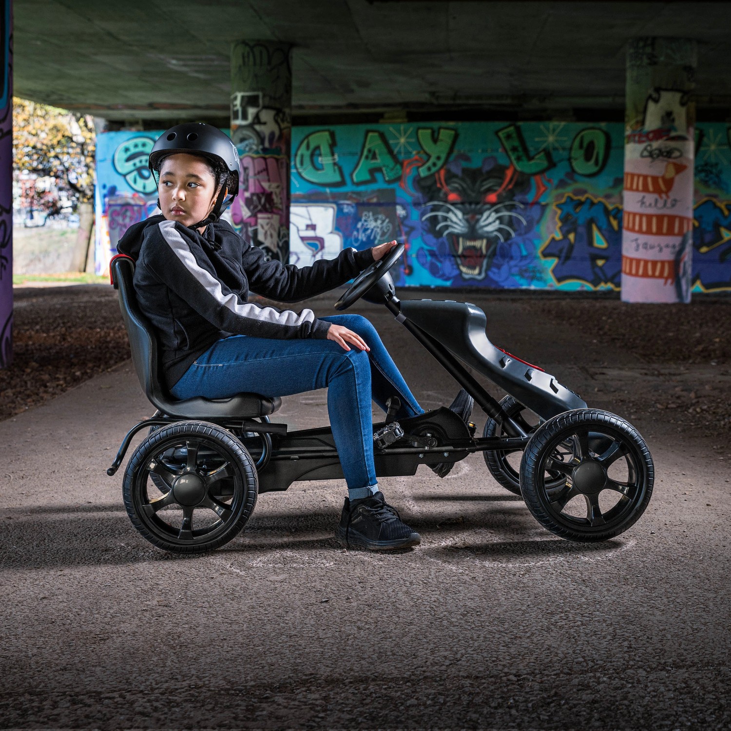 Schwarzes Xootz Go-Kart Venom mit Kind unter Brücke. Das Go-Kart hat profilierte Reifen und einen Schalthebel.