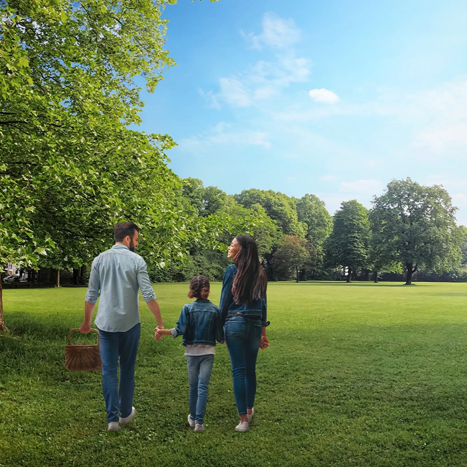 Familie mit Picknickkorb geht durch einen Park mit grüner Wiese und Bäumen.