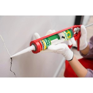 Person dichtet Riss in weißer Wand mit körnigem Soudal Riss & Putz Acryl Weiß aus Kartuschenpistole ab.