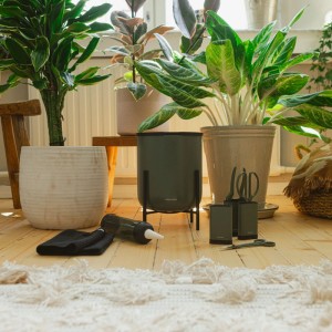 Fiskars Urban Plant Care Selbstbewässerungssystem in Grün mit Zubehör und Pflanzen im Hintergrund.